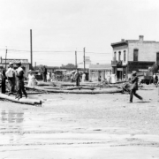 View of a Cherry Creek flood in Denver, Colorado after the Castlewood Canyon Dam break; shows muddy water, the Blake Street bridge, people, debris, and commercial buildings with signs: "American Forge Works."