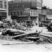 View of Cherry Creek flood damage in Denver, Colorado after the Castlewood Canyon Dam break; shows debris, people, automobiles, office buildings, and signs: "Hotel Revere," "Sharp Bros," and "Standard Machinery & Supply."