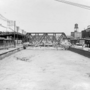 View of Cherry Creek flood damage in Denver, Colorado after the Castlewood Canyon Dam break; shows bridges, muddy water, a water tower, and a warehouse with letters: "Weicker Transfer & Storage Co."