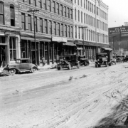 View of Cherry Creek flood damage in Denver, Colorado after the Castlewood Canyon Dam break; shows Wazee Street, mud, automobiles, storefronts, and signs: "Christensen Implement Co," "Oxford Hotel," and "Inland Paper Box Co."