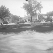 View of the Cherry Creek flood in Denver, Colorado after the Castlewood Canyon Dam break; shows torrents of water, automobiles on Speer Boulevard, and a gas station with tile roof and sign: "Standard Oil Company."