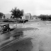 View of a Cherry Creek flood at 8th (Eighth) Avenue and Speer Boulevard in Denver, Colorado. A car is in the foreground; spectators watch by the Nurses Home and County Hospital (Denver General Hospital) in the background.