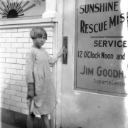 A barefoot girl prepares to open the front door of the Sunshine Rescue Mission at 1822 Larimer Street in Denver, Colorado. The sign in the windows shows a sun with sunbeams that reads: "[Sunshin]e Rescue Mission" and "If [?] Love Shall Conquer Theft." The sign on the door reads: "Hope for all w[?]. Sunshine Rescue Miss[ion]. Service 12 O'clock Noon and [?]. Jim Goodh[eart], Superintende[nt]."