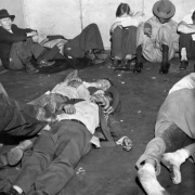 Drunk men sit and lie on the floor of a holding cell in the Police Building, Denver, Colorado. Several men are passed out on the floor. Trash and cigarette butts lie near their bodies.  An African American (Black) man sits with his back against the wall and his head bowed. Two young men sit on either side of him with their arms crossed and heads lowered. A man who sits against the wall smiles a toothless smile.