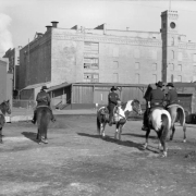 Denver police officers mounted on horses patrol a warehouse district in the Swansea Neighborhood of Denver, Colorado. Shows industrial brick buildings with boarded windows and wooden outbuildings.  A sign near a chain link fence reads: "Peck's Products Company."