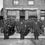 A group of Denver police officers pose near the East Denver Police Station located at 1309 38th (Thirty-eighth) Street in the Five Points Neighborhood of Denver, Colorado. Commanding officers stand in the front row including from left to right: Sergeant Frank Eaton Soward, Captain August Honnebuth, and Sergeant William Ustick. Three motorcycles with side-cars are in the second row.  The remaining police men stand in lines close to the building. Signs on a nearby door read: "1307" and "Rooms For Rent."
