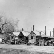 View of makeshift houses and shanties at 19th (Nineteenth) and Clay Streets in the Jefferson Park neighborhood of Denver, Colorado. A man works by barrels and corrugated metal.