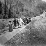 View of a wedding procession in the Colorado mountains; the bride wears a horseshoe, veil, and gown and rides a burro. The groom wears a Civil War uniform; well-dressed people follow.