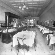 Interior view of the City Cafe restaurant in Denver, Colorado; decor includes a carved wood bar, a tile floor, potted plants, lamps, coving, United States flags, a glass display case of cigars, and a pressed tin ceiling.