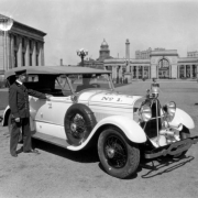 Fire Chief John F. Healy poses in uniform by a Fire Department vehicle (Lincoln sedan) in Civic Center park, Denver, Colorado. The automobile has a bell and siren. The Voorhies Memorial Colonnade, Carnegie Library, and Arapahoe County Courthouse dome are in the background.