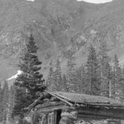 A miner, in a striped suit with a pick axe, poses next to a log cabin in possibly Clear Creek County, Colorado. The cabin has a gabled roof, a metal chimney, a dirt roof and a sign in a window that reads: "[C]rocker [& Co.] Crac[kers], Denver." Trees are near the cabin, steep slopes and talus are on mountains above timberline.