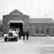 African American (Black) and white firemen pose by a fire truck and Station No. 3 of the Denver Fire Department at 2363 Glenarm Place in the Curtis Park (Five Points) neighborhood of Denver, Colorado. The men wear uniforms and caps. The station has brick arches, keystones, and a tile roof.