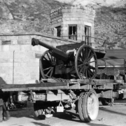 Chained to a flatbed truck at the State Penitentiary in Canon City, Colorado, is a wheel-mounted artillery, 75 millimeter field piece from the National Guard firing range in Golden. It was used during the October 3 riot after dynamite failed to roust the ringleaders. A man in gaiters moves metal ammunition boxes. Windows in the stone wall and guard tower are broken.