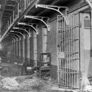 Interior, bracketed metal and pipe railed balconies top cells at the State Penitentiary in Canon City, Colorado. Iron gates are in the brick wall; one in the foreground is open. A writing slate hangs inside the cell, and the facing floor is strewn with blood, debris and a wooden chair. The row-lock is by the door, and a fallen beam leans on the balcony.