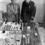 Tools used in a prison break from the State Penitentiary in Canon City, Colorado, include hacksaw blades, drill brace and bits, pliers, a screwdriver, putty knife, batteries, string, wire, and tape. In a Fedora, tweed jacket and tie, associate warden Fred Wyse holds a serrated knife as Captain Chester Yeo, in uniform, looks on. Also on the table are gloves, a spool of fishing line, and Colorado license plate number: "KD- 4989," 1962.