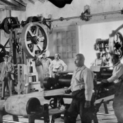 Inmates (one Black) and officials at the State Penitentiary in Canon City, Colorado, pose among metalworking machines with rollers, flywheels and belted pulleys. An axle mounted on an interior wall drives a drill press and a stamp mill, which has "US Toledo No. 57" embossed on its frame.  Track mounted, sliding wooden doors with paned windows are open, and glare comes in from outside. The men are in striped shirts, coveralls, and a suit and tie.