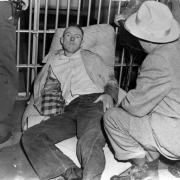 Lying on a wadded, pinstriped mattress, This captured escapee from the State Penitentiary in Canon City, Colorado, appears defiant in his dungarees, shirt and stained jacket. Interior prison bars are behind him; the man squatting in the foreground is identified as Red Fenwick.