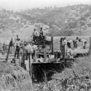 Men work at the State Penitentiary in Canon City, Colorado. Plank walls and roof surround an opening fronted by a channel in the foreground. With bags, shovels, and wheelbarrows, inmates are watched by men sitting on barrels in a mule drawn wagon. Power lines, railroad track, and low hills are in the background.