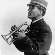 Cornet in hands, the leader of the State Penitentiary Band in Canon City, Colorado poses in profile. His uniform has appliqued braiding, and his cap has a shiny bill and brass band insignia. He wears wire rimmed spectacles.