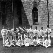 Members of the prisoner's band at the State Penitentiary in Canon City, Colorado, pose with drums, trombones, cornets, and trumpets. Their uniforms are white with rectangles across the fronts, and they wear conductor's hats. Stone Prison buildings with extruded joints and arched, barred windows are behind them.