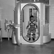 A man poses strapped to a chair in the gas chamber at the State Penitentiary in Canon City, Colorado. He wears pants, belt and shoes; a tube connected to his chest is linked to a stethoscope under a window on the exterior of the unit. The door has a wheel closure; adjacent pipes are topped with wheel valves. A lever and spring are in back, a white plastic jar is atop the door mechanism.