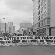 Mexican Americans, members of the Crusade for Justice (La Crusada Para Justicia) march in protest down 15th (Fifteenth) street in Denver, Colorado. They carry a large banner written in both English and Spanish, which reads: "It Is Better to Die On Your Feet Than to Keep On Living On Your Knees," "Es Mejor Morir de Pie, Que Seguir Viviendo de Rodillas."