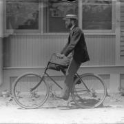 A man in a suit and straw hat poses on a bicycle in (probably) Denver, Colorado. He wears spectacles and a beard; a leather case is strapped to the bike.