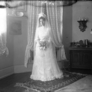 Blanche Roberts poses in possibly a wedding dress with a veil, in (probably) Denver, Colorado. She holds a bouquet of flowers; another woman sits to the side. Decor includes a woven rug and a sconce shelf with a mounted taxidermic antelope and antlers.
