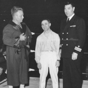 Men stand and pose in a boxing ring. Joe Rutofsky "Awful" Coffee smiles and poses between two men. Everett O. Marshall wears lace-up boxing shoes and a robe with satin lapels, cuffs, and a belt. William Harrison (Jack) Dempsey wears a military uniform with a double breasted jacket and three stripes on the sleeves.