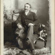 Portrait of Dr. John Elsner. He has a moustache and wears a shirt with a starched collar, probably a silk band bow tie, a double breasted jacket with probably satin lapels, pin-striped slacks, and leather boots with covered laces. Dr. Elsner sits on fur pelts on a carved wooden armchair with floral upholstery. Fur pelts on the ground include a coyote or fox pelt.