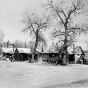 View of shanties on a dirt road near probably 19th (Nineteenth) Avenue and Clay Street in the Jefferson Park neighborhood of Denver, Colorado. Houses are made of wood and corrugated metal and have chimneys and roofs patched with shingles and sheet metal. A man in the road wears a v-neck sweater and a hat and smokes a pipe. Trees, scrap wood, metal pots, and laundry on a clothesline are beside buildings.