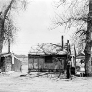 A man stands with arms crossed in front of a shanty near probably 19th (Nineteenth) Avenue and Clay Street in the Jefferson Park neighborhood of Denver, Colorado. Houses are made of wood and corrugated metal with metal chimneys. A house has a scalloped edge over the eves of a gabled roof. Pots and pans hang by a window, and laundry, wooden trunks, and metal buckets are near trees.