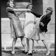 Margie, June and Charlotte, three teenage African American girls, pose in front of the home of Virginia Woodward, their Sunday School teacher, in Denver, Colorado. They wear dresses and stockings; one wears a jacket; they stand in a circle and hold hands.