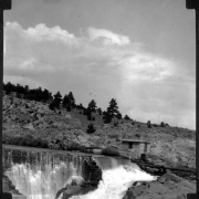 View of the dam at the entrance of Big Thompson Canyon (Larimer County), Colorado. Water flows over the dam, and a small concrete building near large pipes is shown.