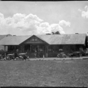 View of the Administration Building at the Y.M.C.A. Camp Conference Headquarters at the YMCA of the Rockies near Estes Park in Larimer County, Colorado. Shows men, women and children in front of a two-story, wood frame building. Cars nearby include Model "A" Fords.
