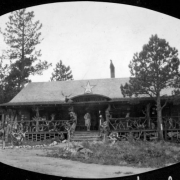 View of the souvenir shop on a fox farm in Boulder County, Colorado. Shows a wood frame building with a porch railing made of tree branches. A man and a woman stand on the porch. A woman walks away from the porch. Signs over the doorway read: "Dolley's," and "Welcome."