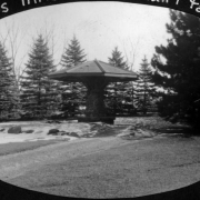 View of the sun shelter at the Sunken Gardens near West High School in Denver, Colorado. Shows a pillar made of large stones, stone bench and wooden roof. Spruce and pine trees are in the background.