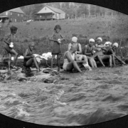 A group of African American campers from Camp Nizhoni bathe on the bank of South Boulder Creek in Lincoln Hills (Gilpin County), Colorado. The teenage girls wash clothes and wear swim suits and caps. Camp buildings are in the distance.