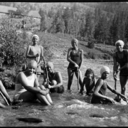 Pictured are Declora James, Zudora Blakey, Mary E. Wood ("Woody," the Camp's Director), Marie Anderson, a camp counselor (later Marie L. Greenwood), Ruby Hawkins, Ethel Wood, Marie Yarbra, and Eleanor Hawkins ("Hawky, " a camp counselor) African American females from Camp Nizhoni in Lincoln Hills (Gilpin County), Colorado. They stand and sit in the South Boulder Creek while wearing swim suits and swim caps; some hold large sticks.