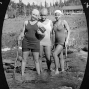 Three African American camp counselors identified as: "Smitty" (Eleanor Smith), "Woody" (Mary E. Wood) and "Hawky" (Eleanor Hawkins) stand in South Boulder Creek wearing swim suits and swim caps on the grounds of Camp Nizhoni in Lincoln Hills (Gilpin County), Colorado. "Smitty" leans on a large stick and holds a box camera; "Hawky" also leans on a large stick.