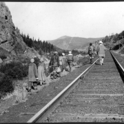 African American teenage campers and their counselor from Camp Nizhoni in Lincoln Hills hike along Denver & Rio Grande Western Railroad tracks near Rollinsville (Gilpin County), Colorado. The buildings of the town are in the distance. The hikers wear coats, sweaters, pants, boots and hats. One carries a box camera; another carries a cup.