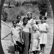 Six African American campers of various ages in the "Andy" family (Andy is counselor Marie Anderson, later Marie L. Greenwood) pose near a wire fence on the grounds of Camp Nizhoni in Lincoln Hills (Gilpin County), Colorado. Some wear dresses, two wear hats, and one wears a long skirt. Camp buildings are in the distance.