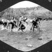 African American teenage campers wear swim suits and swim caps as they splash in South Boulder Creek in Camp Nizhoni in Lincoln Hills (Gilpin County), Colorado. A campground building and flagpole are in the distance.
