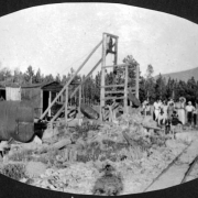 View of the headframe of a tungsten mine and large metal tanks. African American female campers and counselors from Camp Nizhoni stand by the mine, probably in Gilpin County, Colorado. Shows ore car tracks.