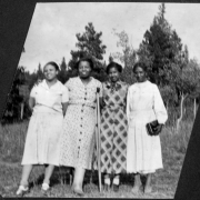 Four African American women, Katherine Patton (Pat), Mary E. Wood (Woody), Marie Anderson ( Andy, later Marie L. Greenwood), and Mrs. Wilson pose together at Camp Nizhoni in Lincoln Hills (Gilpin County), Colorado. Woody has a broken leg and leans on one crutch. They all wear dresses; two of which have a print pattern. Mrs. Wilson holds a purse.