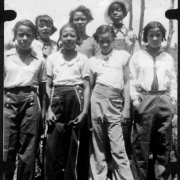 Seven African American teenage campers from Camp Nizhoni pose in Lincoln Hills (Gilpin County), Colorado. They all wear pants and blouses; one has on overalls, one wears a straw hat and three others wear sailor-type pants with large buttons.