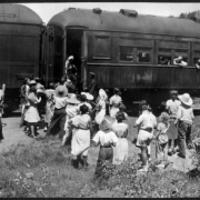 Passengers look out windows as a group of African American campers and their counselors from Camp Nizhoni (Gilpin County), Colorado stand near a D.& R.G.W. Railroad train near Lincoln Hills. The campers and counselors wear pants, shorts, dresses and wide-brimmed hats. The train car reads: "Railway Express Agency Baggage 561."
