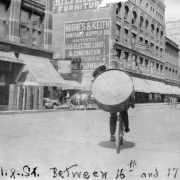 View of California Street in Denver, Colorado; a bicycle rider carries a bass drum. Horse-drawn wagons are by office buildings and storefronts with signs that read: "Hughes & Keith Sanitary Supply Co." and "R. Douglas Crockery Co."