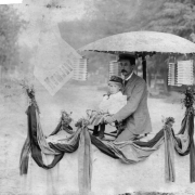 A man and child ride a bicycle decorated with crepe paper, an umbrella, and Japanese lanterns, in probably Denver, Colorado.