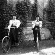 Women pose on bicycles in Denver, Colorado; outfits include straw hats, puff sleeved blouses, gloves, and a purse. A brick and stone building is covered in vines.